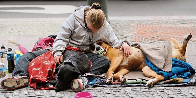 Ein Frau sitzt mit zwei Hunden an einem Laternenpfahl auf dem Gehweg