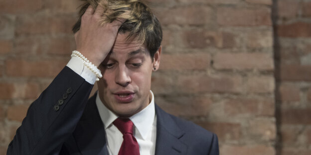 Ein Mann im Anzug fährt sich mit rechten Hand durch die Haare. Es ist Milo Yiannopoulos Ein Mann im Anzug fährt sich mit rechten Hand durch die Haare. Es ist Milo Yiannopoulos