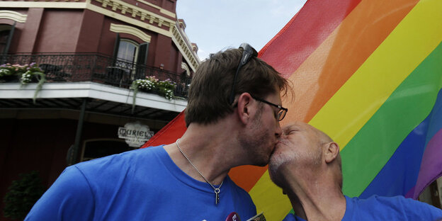 Zwei Männer, beide in blauen T-Shorts, küssen sich vor einer Regenbogen-Flagge Zwei Männer, beide in blauen T-Shorts, küssen sich vor einer Regenbogen-Flagge