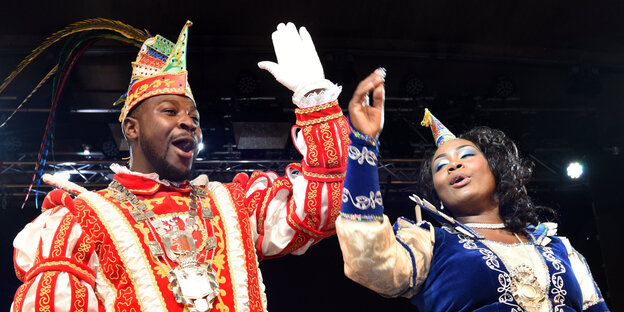 Samuel I. und Jacinta I. stehen in Karnevals-Kluft in einem Festsaal. Sie heben die Arme und singen Samuel I. und Jacinta I. stehen in Karnevals-Kluft in einem Festsaal. Sie heben die Arme und singen