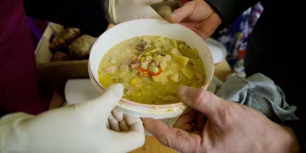 Jemand überreicht einer anderen Person einen Plastikteller voll Suppe Jemand überreicht einer anderen Person einen Plastikteller voll Suppe