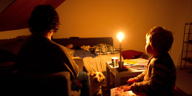 Eine Frau und ein Kind sitzen an einem Wohnzimmertisch. Der Raum wird nur durch eine Kerze erhellt