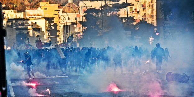 Im Hintergrund die Fassaden mehrerer Häuser in Neapel, im Vordergrund eine Menschenmenge vor denen es raucht und kleine Flammen brennen Im Hintergrund die Fassaden mehrerer Häuser in Neapel, im Vordergrund eine Menschenmenge vor denen es raucht und kleine Flammen brennen