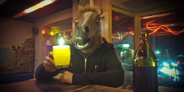 Ein Mensch mit Pferdemaske sitzt an einem Tresen und prostet mit Bier in die Kamera