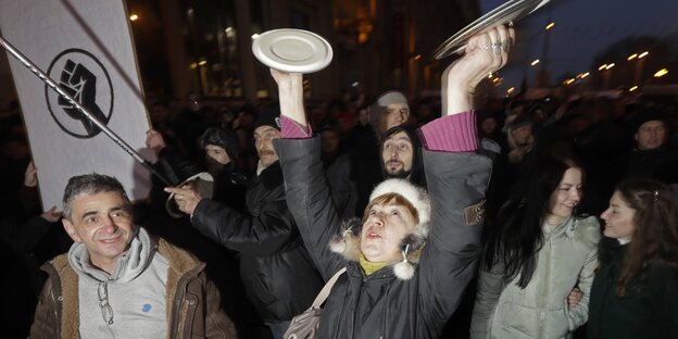 Eine Frau schlägt in einer Menge von Demonstranten zwei Topfdeckel gegeneinander Eine Frau schlägt in einer Menge von Demonstranten zwei Topfdeckel gegeneinander