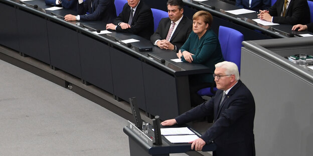 Ein Mann mit weißen Haaren steht am Rednerpult, weitere Menschen sitzen im Hintergrund und hören ihm zu Ein Mann mit weißen Haaren steht am Rednerpult, weitere Menschen sitzen im Hintergrund und hören ihm zu
