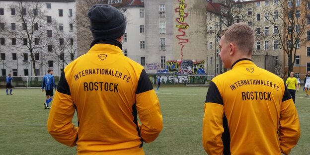 Zwei Männer stehen mit dem Rücken zur Kamera und blicken auf ein Fußballspiel. Auf ihren Jacken steht „Internationaler FC Rostock“ Zwei Männer stehen mit dem Rücken zur Kamera und blicken auf ein Fußballspiel. Auf ihren Jacken steht „Internationaler FC Rostock“