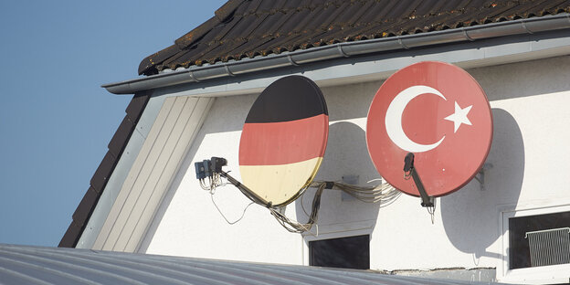 An einer Hauswand unter einem Giebel sind zwei Satellitenschüsseln installiert. Eine ist mit den Farben der deutschen Fahne bemalt, die andere mit den Farben der türkischen An einer Hauswand unter einem Giebel sind zwei Satellitenschüsseln installiert. Eine ist mit den Farben der deutschen Fahne bemalt, die andere mit den Farben der türkischen