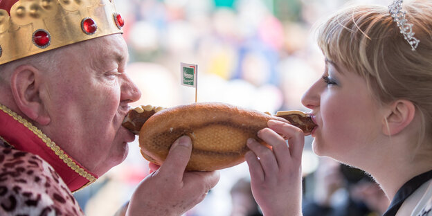 Links ein alter Mann mit Krone, rechts eine junge Frau mit Diadem. Sie beißen gemeinsam in eine Bratwurstsemmel, die in der Bildmitte ist Links ein alter Mann mit Krone, rechts eine junge Frau mit Diadem. Sie beißen gemeinsam in eine Bratwurstsemmel, die in der Bildmitte ist