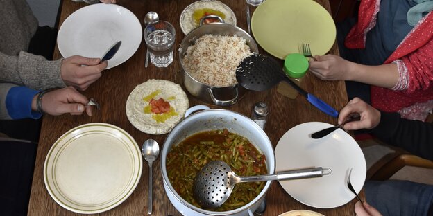 Das Essen ist fertig und steht auf dem Tisch, an dem Bilal und drei Freunde von ihm sitzen. Der Tisch, ein Holztisch, ist komplett voll. Darauf steht ein Topf mit Aldi b'set, den grünen Bohnen, ein Topf mit Reis, ein Salat und Hummus-Creme. Das Essen ist fertig und steht auf dem Tisch, an dem Bilal und drei Freunde von ihm sitzen. Der Tisch, ein Holztisch, ist komplett voll. Darauf steht ein Topf mit Aldi b'set, den grünen Bohnen, ein Topf mit Reis, ein Salat und Hummus-Creme.