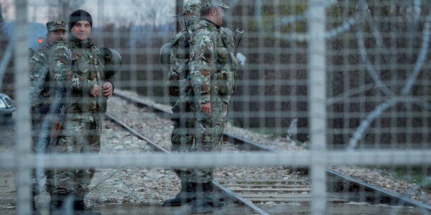 Im Vordergrund Zaun mit Stacheldraht, im Hintergrund Soldaten