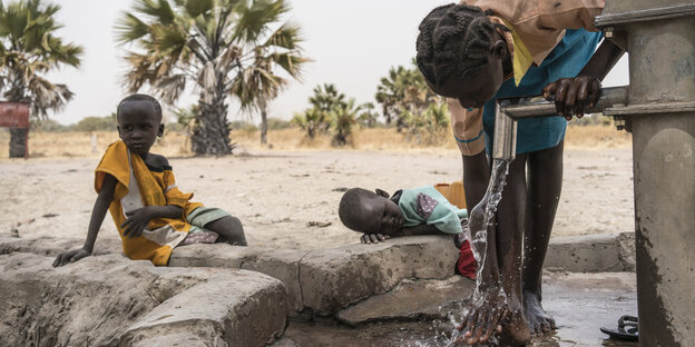 Eine Frau wäscht ihre Füße an einem Brunnen. Daneben sitzen und liegen zwei Kinder und schauen ihr zu Eine Frau wäscht ihre Füße an einem Brunnen. Daneben sitzen und liegen zwei Kinder und schauen ihr zu
