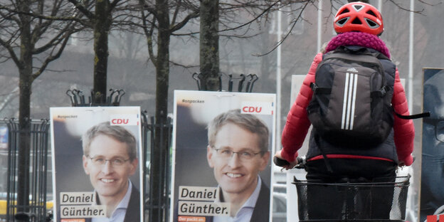 Links im Bild zwei Wahlplakate mit dem Porträtfoto eines Mannes, rechts im Bild eine Fahrradfahrerin mit Helm von hinten Links im Bild zwei Wahlplakate mit dem Porträtfoto eines Mannes, rechts im Bild eine Fahrradfahrerin mit Helm von hinten