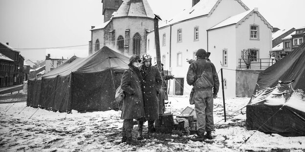 Ein schwarz-weiß Foto zeigt eine Gruppe Soldaten, im Hintergrund zwei Zelte und Häuser eines Dorfs Ein schwarz-weiß Foto zeigt eine Gruppe Soldaten, im Hintergrund zwei Zelte und Häuser eines Dorfs