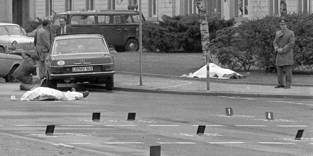 Schwarzweißfoto: Straße mit Polizeimarkierungen, stehenden Fahrzeugen und zwei zugedeckten Körpern Schwarzweißfoto: Straße mit Polizeimarkierungen, stehenden Fahrzeugen und zwei zugedeckten Körpern