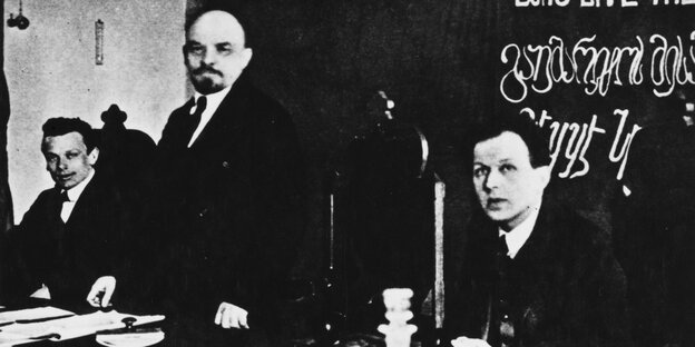 Ein schwarz-weiß Bild: Drei Männer nebeneinander, die beiden äußeren sitzen, der in der Mitte steht an einem Tisch Ein schwarz-weiß Bild: Drei Männer nebeneinander, die beiden äußeren sitzen, der in der Mitte steht an einem Tisch