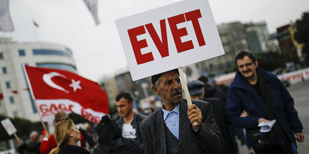 Im Vordergrund ist ein älterer Mann mit einem Schild auf dem "Evet", also "Ja" steht, im Hintergrund stehen weitere Menschen, einer schwingt eine türkische Flagge Im Vordergrund ist ein älterer Mann mit einem Schild auf dem "Evet", also "Ja" steht, im Hintergrund stehen weitere Menschen, einer schwingt eine türkische Flagge