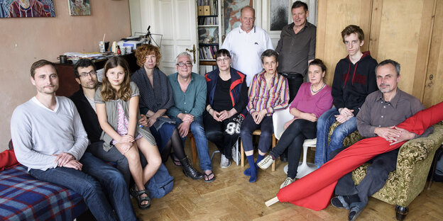 Eine Gruppe von Menschen sitzt im Zimmer einer Wohnung auf Stühlen und einem Bett Eine Gruppe von Menschen sitzt im Zimmer einer Wohnung auf Stühlen und einem Bett