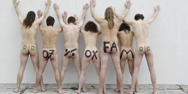 Sieben nackte Menschen stehen an einer weißen Wand, mit dem Rücken zum Betrachter. Auf ihren Hintern steht gemalt: 0,7% Oxfam Sieben nackte Menschen stehen an einer weißen Wand, mit dem Rücken zum Betrachter. Auf ihren Hintern steht gemalt: 0,7% Oxfam