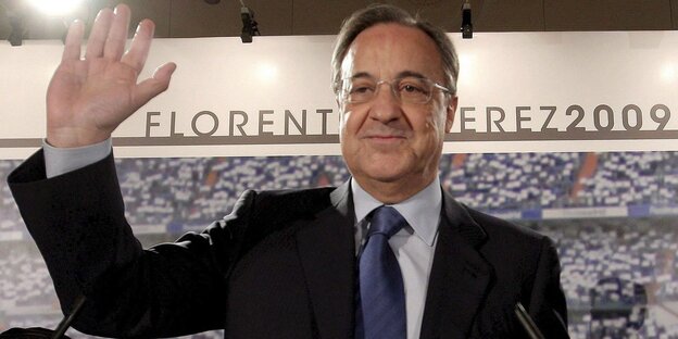 Ein lächelnder winkender Mann in Anzug und Krawatte vor den Rängen eines Stadions und dem Schriftzug Florentino Pérez. Ein lächelnder winkender Mann in Anzug und Krawatte vor den Rängen eines Stadions und dem Schriftzug Florentino Pérez.