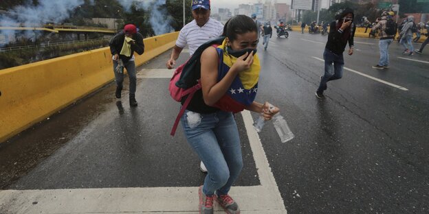 Eine Gruppe Demonstranten läuft auf einer Straße auf den Betrachter zu. Sie werden mit Tränengas beschossen. Im Vordergrund eine Frau. Sie hält zwei Flaschen in der linken Hand und bedeckt ihren Mund mit einer venezualischen Fahne. Eine Gruppe Demonstranten läuft auf einer Straße auf den Betrachter zu. Sie werden mit Tränengas beschossen. Im Vordergrund eine Frau. Sie hält zwei Flaschen in der linken Hand und bedeckt ihren Mund mit einer venezualischen Fahne.