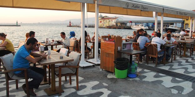 Menschen sitzen im Freien in der Abendsonne an Tischen in einem Café am Wasser. Menschen sitzen im Freien in der Abendsonne an Tischen in einem Café am Wasser.