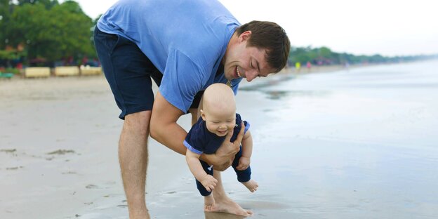 Ein Vater steht barfuß am Strand, im Arm hält er ein kleines Kind. Er beugt sich vor, um dem Kind den Sand zu zeigen und lacht Ein Vater steht barfuß am Strand, im Arm hält er ein kleines Kind. Er beugt sich vor, um dem Kind den Sand zu zeigen und lacht