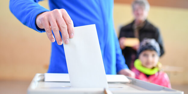 Eine Person in blauem Pulli steckt einen Wahlzettel in eine Wahlurne. Eine Person in blauem Pulli steckt einen Wahlzettel in eine Wahlurne.