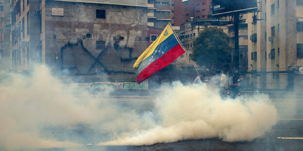 gelblicher Rauch von Tränengas zieht über eine Straße in Caracas, mittendrin halten Demonstranten die venezolanische Flagge hoch gelblicher Rauch von Tränengas zieht über eine Straße in Caracas, mittendrin halten Demonstranten die venezolanische Flagge hoch