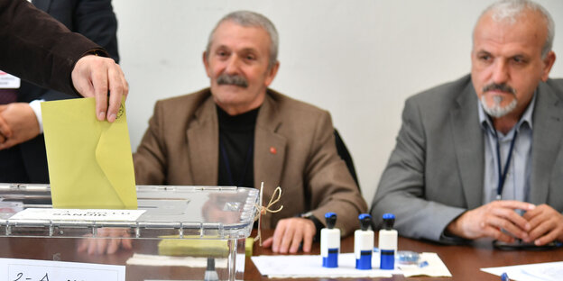 Eine Hand wirft einen gelben Umschlag in eine Plastikbox, im Hintergrund sehen zwei ältere Männer dabei zu Eine Hand wirft einen gelben Umschlag in eine Plastikbox, im Hintergrund sehen zwei ältere Männer dabei zu