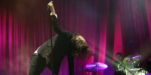 Father John Misty steht auf der Bühne. Er hält sich mit einer Hand am Mikroständer fest und biegt sich rücklings nach hinten, sein Arm und seine langen Haare hängen nach unten