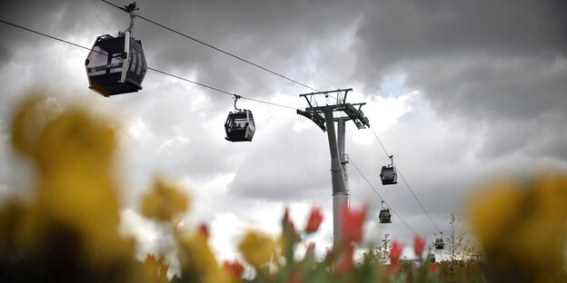 Seilbahngondeln vor einem wolkenverhangenen Himmel, verschwommen im Vordergrund: Osterglocken Seilbahngondeln vor einem wolkenverhangenen Himmel, verschwommen im Vordergrund: Osterglocken
