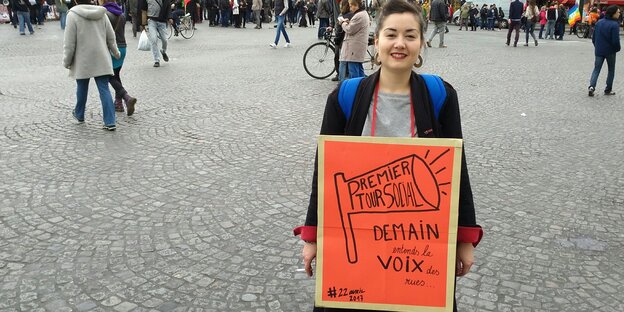 Eine Frau mit einem orangefarbenen Papschild steht auf einem Platz Eine Frau mit einem orangefarbenen Papschild steht auf einem Platz