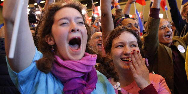 Zwei Frauen, eine im hellblauen, eine im rosa Macron-T-Shirt jubeln, strahlen und eine schwenkt ein Fähnchen