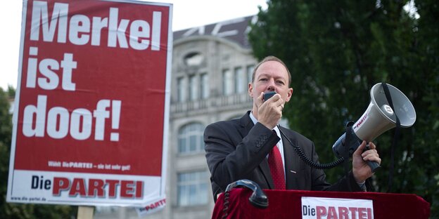 Ein Mann hält ein Plakat mit der Aufschrift "Merkel ist dick!" in die Höhe, daneben sieht man weitere Plakate der Partei die Partei