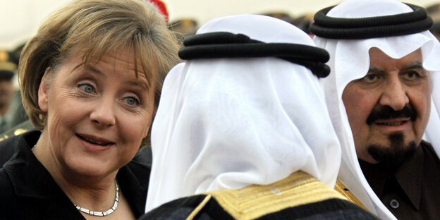 Angela Merkel lacht, neben ihr steht ein Mann mit traditioneller Kopfbedeckung Angela Merkel lacht, neben ihr steht ein Mann mit traditioneller Kopfbedeckung