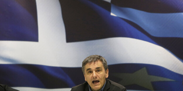 Der griechische Finanzminister Euklid Tsakalotos sitzt auf einer Pressekonferenz vor einer großen griechischen Flagge Der griechische Finanzminister Euklid Tsakalotos sitzt auf einer Pressekonferenz vor einer großen griechischen Flagge