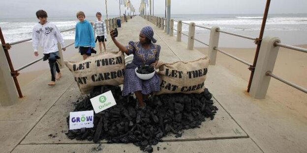 Aktivistin auf einem Kohlehaufen auf einer Seebrücke, zwei Säcke tragen die Aufschrift: "Let them eat carbon“ Aktivistin auf einem Kohlehaufen auf einer Seebrücke, zwei Säcke tragen die Aufschrift: "Let them eat carbon“
