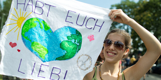 Eine junge Frau mit Sonnenbrille hält ein Stück Stoff mit einem darauf gemalten Herz neben ihr Gesicht