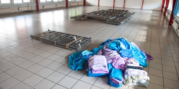 In einer großen Halle stehen zusammengelegte Bettgestelle und Wäsche In einer großen Halle stehen zusammengelegte Bettgestelle und Wäsche