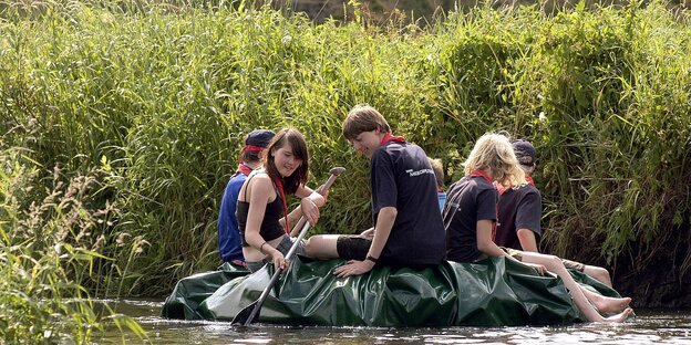 Mehrere Jugendliche paddeln in einem Schlauchboot an einem Schlifufer entlang