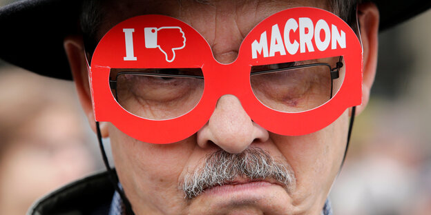 Ein Mann trägt eine rote Pappbrille, auf der ein Dislike-Symbol und Macron zu sehen sind Ein Mann trägt eine rote Pappbrille, auf der ein Dislike-Symbol und Macron zu sehen sind