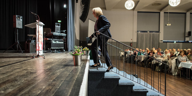 In einem Saal, in dem Zuschauer an Tischen sitzen, geht eine Frau über einige Treppenstufen auf eine Bühne.