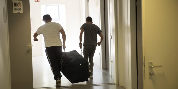 Zwei Jungen tragen gemeinsam eine Tasche einen Flur entlang, auf den Ausgang zu Zwei Jungen tragen gemeinsam eine Tasche einen Flur entlang, auf den Ausgang zu