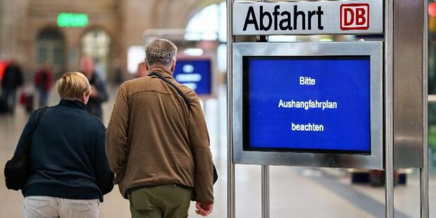 Anzeigedisplay mit dem Hinweis „Bitte Aushangfahrplan beachten“ in einem Bahnhofsgebäude, dahinter läuft ein Pärchen