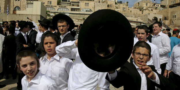 Menschen in orthodox-jüdischer Kleidung in Jerusalem. Im Vordergrund Kinder, ein Junge hält einen schwarzen Hut in die Kamera Menschen in orthodox-jüdischer Kleidung in Jerusalem. Im Vordergrund Kinder, ein Junge hält einen schwarzen Hut in die Kamera