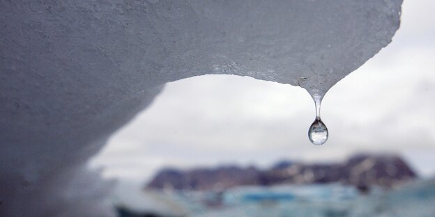 An einem schmelzenden Eisberg hängt ein Tropfen Wasser, im Hintergrund sieht man eisige Landschaften An einem schmelzenden Eisberg hängt ein Tropfen Wasser, im Hintergrund sieht man eisige Landschaften
