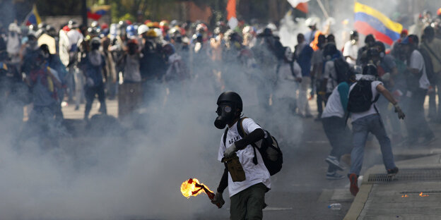 Ein Demonstrant mit Gasmaske vor dem Gesicht hält eine entzündete Flaschenbombe in der Hand. Hinter ihm ist eine Demonstration zu sehen. Rauch verdeckt die Sicht etwas. Ein Demonstrant mit Gasmaske vor dem Gesicht hält eine entzündete Flaschenbombe in der Hand. Hinter ihm ist eine Demonstration zu sehen. Rauch verdeckt die Sicht etwas.
