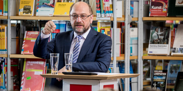 Ein glatzköpfiger Redner an einem Podium mit SPD-Logo vor Bücherregalen. Im Vordergrund die Köpfe einer Zuhörergruppe. Ein glatzköpfiger Redner an einem Podium mit SPD-Logo vor Bücherregalen. Im Vordergrund die Köpfe einer Zuhörergruppe.