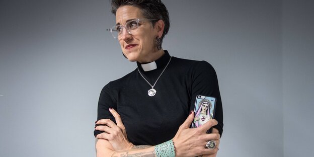 Eine Pastorin steht mit verschränkten Armen da und lächelt, ihr Smartphone trägt ein Marienbild Eine Pastorin steht mit verschränkten Armen da und lächelt, ihr Smartphone trägt ein Marienbild